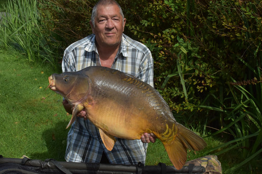 Photo of Ken Townley holding a large carp on the riverside in a check shirt