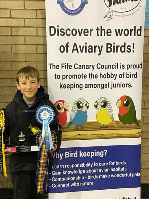 Boy holding a winning cage with rosettes, next to a Haith's banner. 